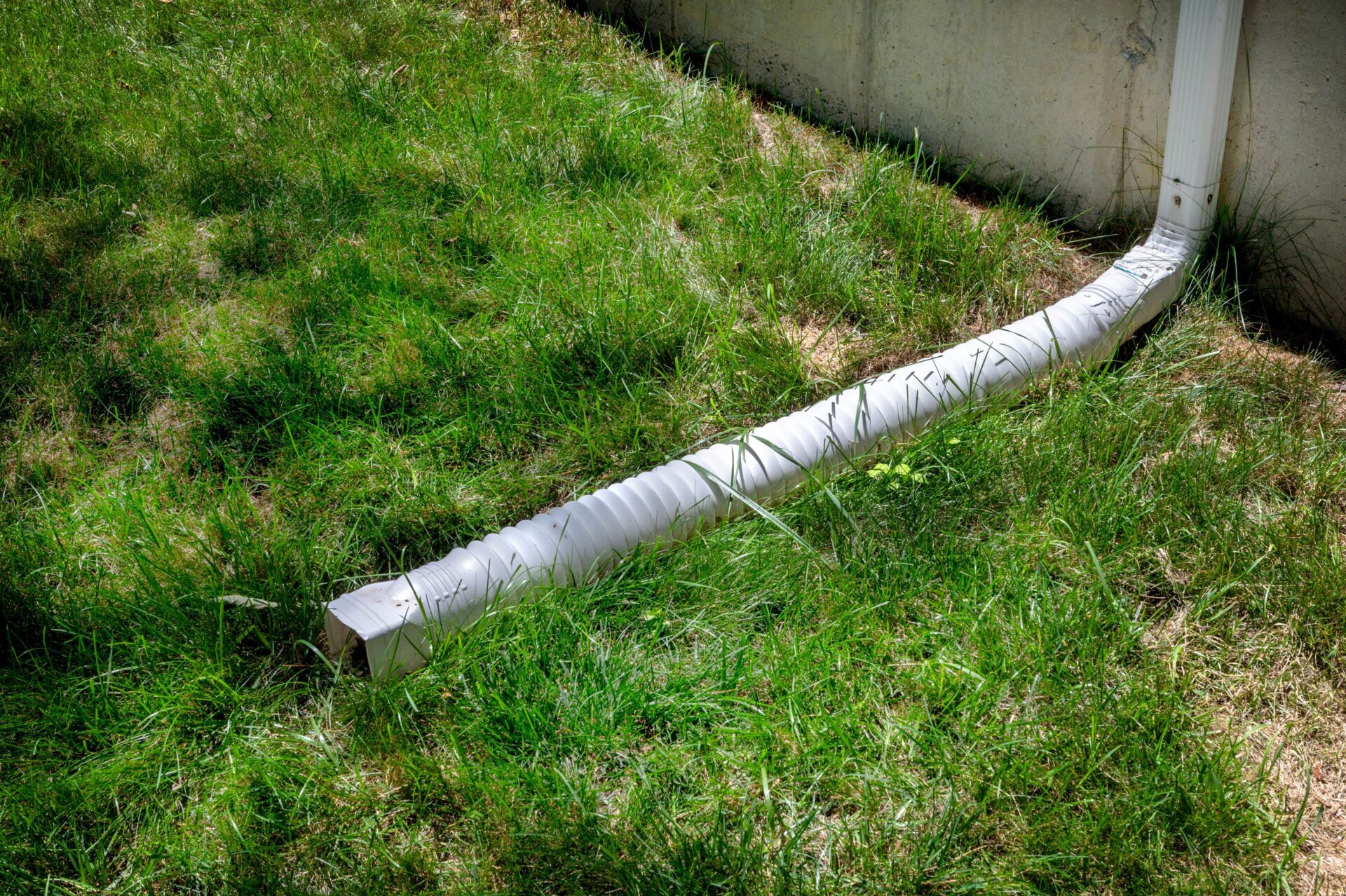 A black plastic downspout extension attached to a white downspout, directing rainwater away from a home foundation onto a grassy yard