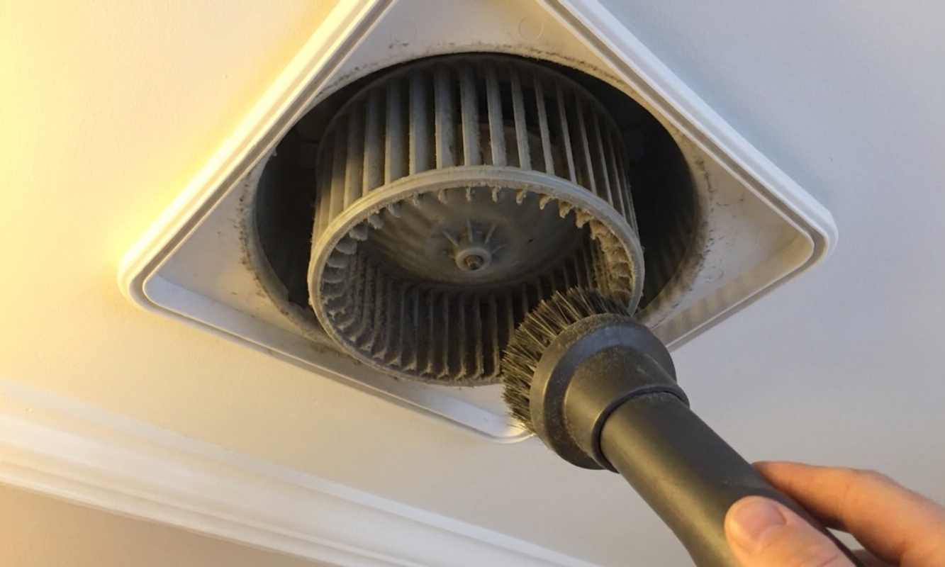 A close-up of a bathroom exhaust fan blower wheel being cleaned with a vacuum brush attachment while the grille is removed