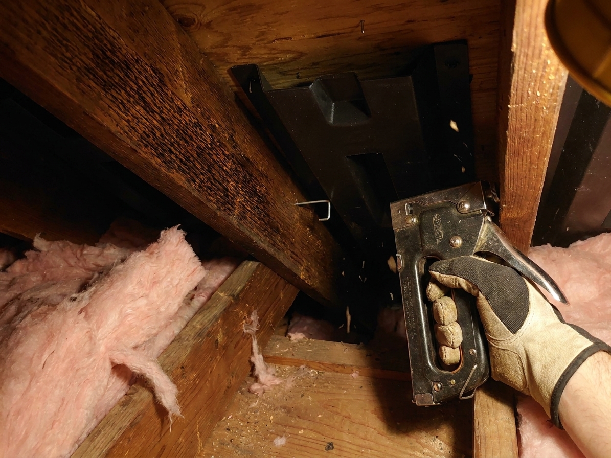 A close-up photo of a hand using a staple gun to fasten a plastic attic ventilation baffle to a wooden rafter inside an attic