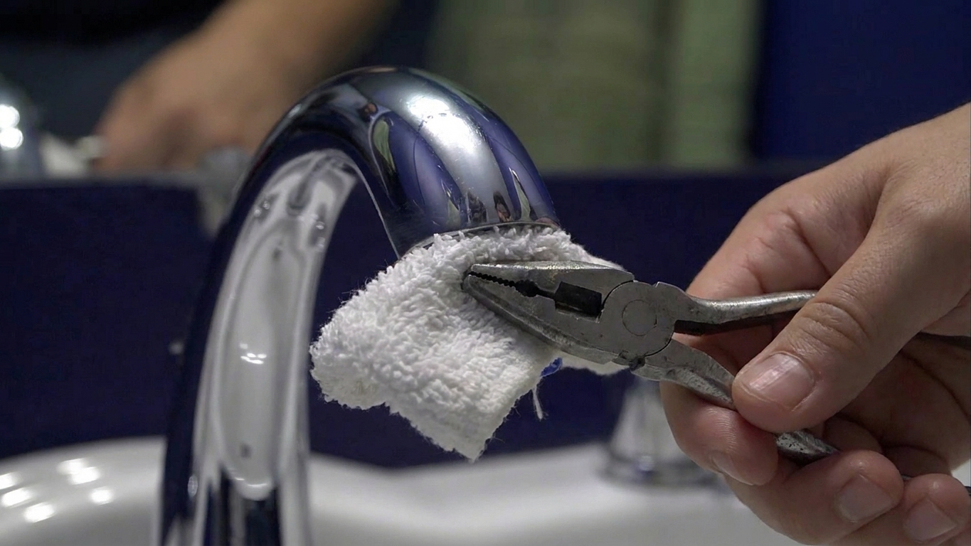 A close-up photo of pliers gripping a towel-wrapped faucet aerator at the end of a chrome bathroom faucet