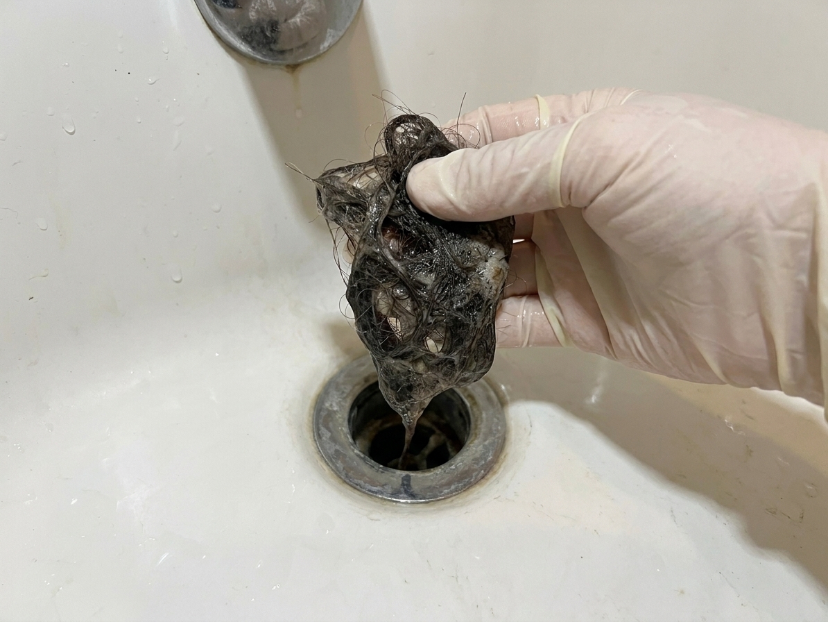 A gloved hand holding a clump of hair and soap scum pulled from a bathtub drain opening