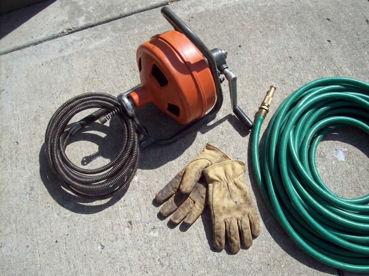 A hand crank drain snake (plumber’s auger) resting on a driveway next to work gloves and a garden hose