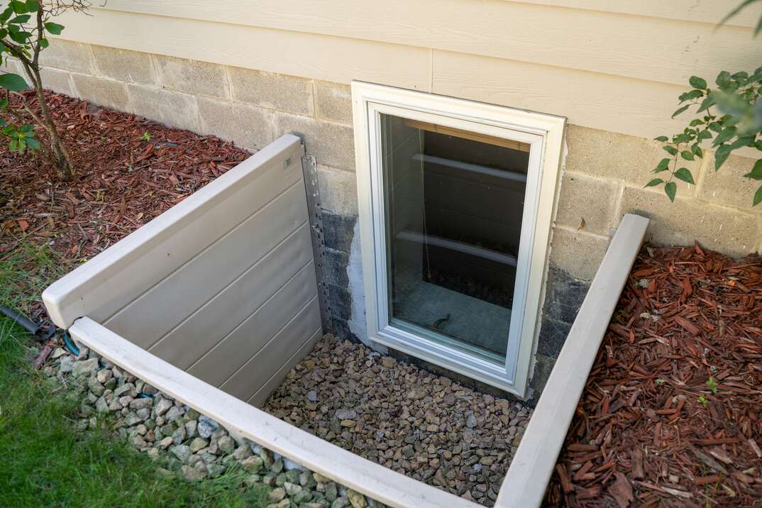 A homeowner clearing wet leaves out of a metal window well next to a basement window, with a small hand rake and bucket nearby