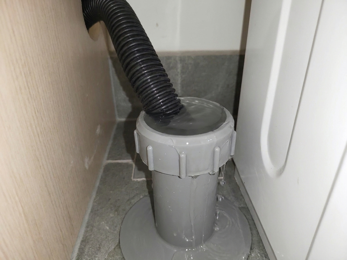 A laundry standpipe behind a washing machine with water pooled at the top edge of the pipe, suggesting it is close to overflowing during a drain cycle