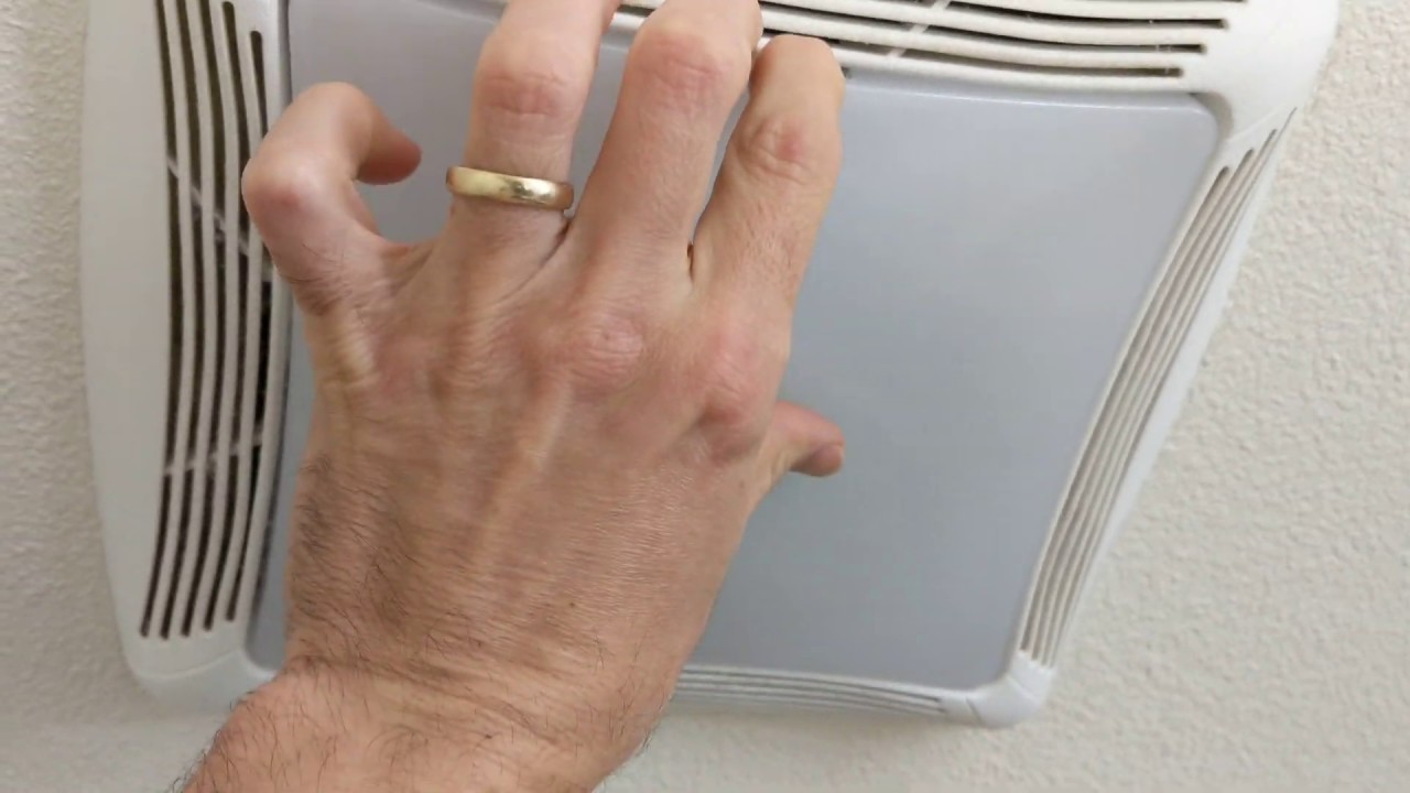 A real bathroom exhaust fan grille partially unclipped from the ceiling, showing dust buildup on the louvers and the spring clips