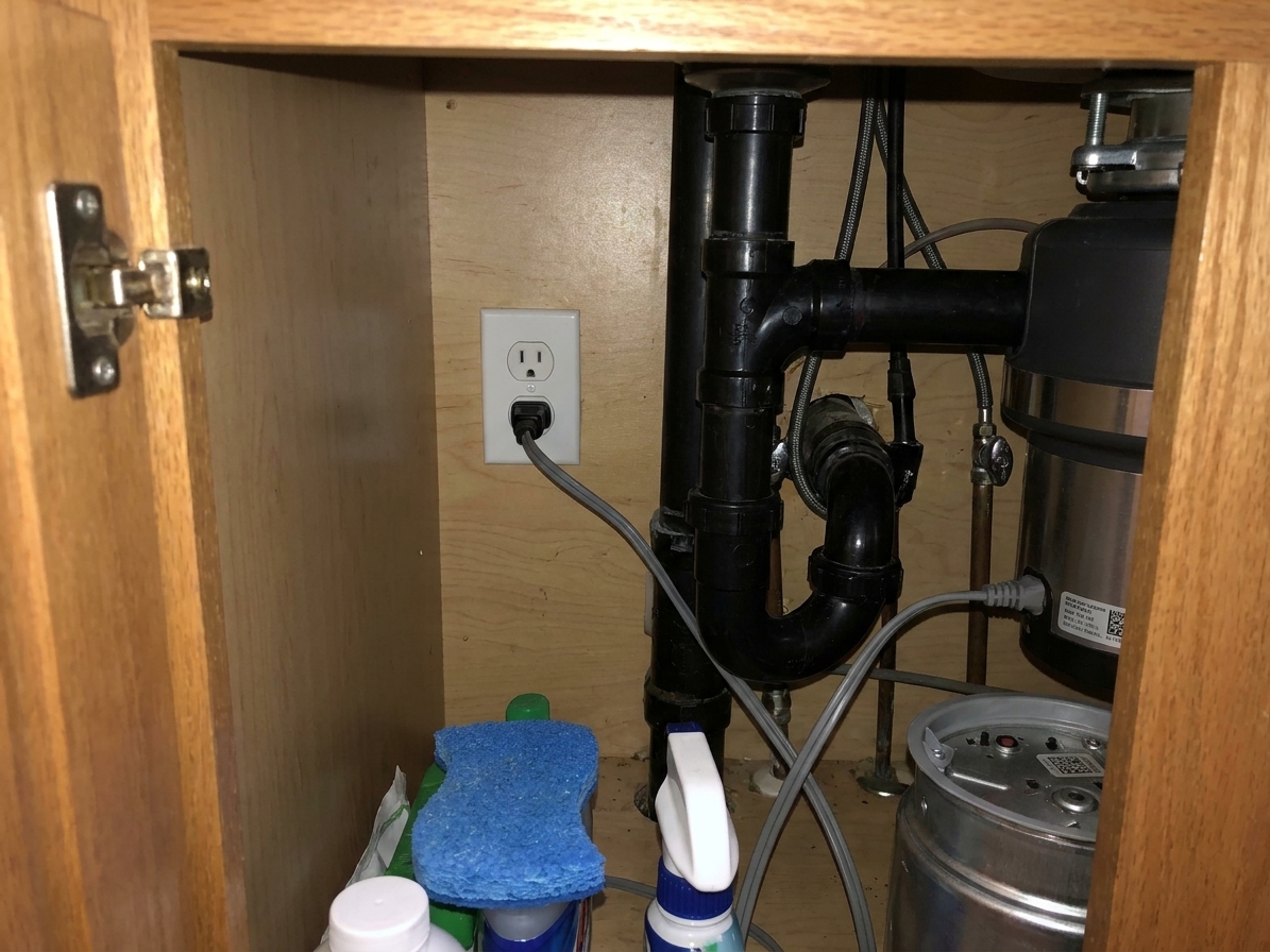 A real-life photo inside a kitchen sink base cabinet showing a standard electrical outlet mounted to the cabinet wall with a disposal plug nearby