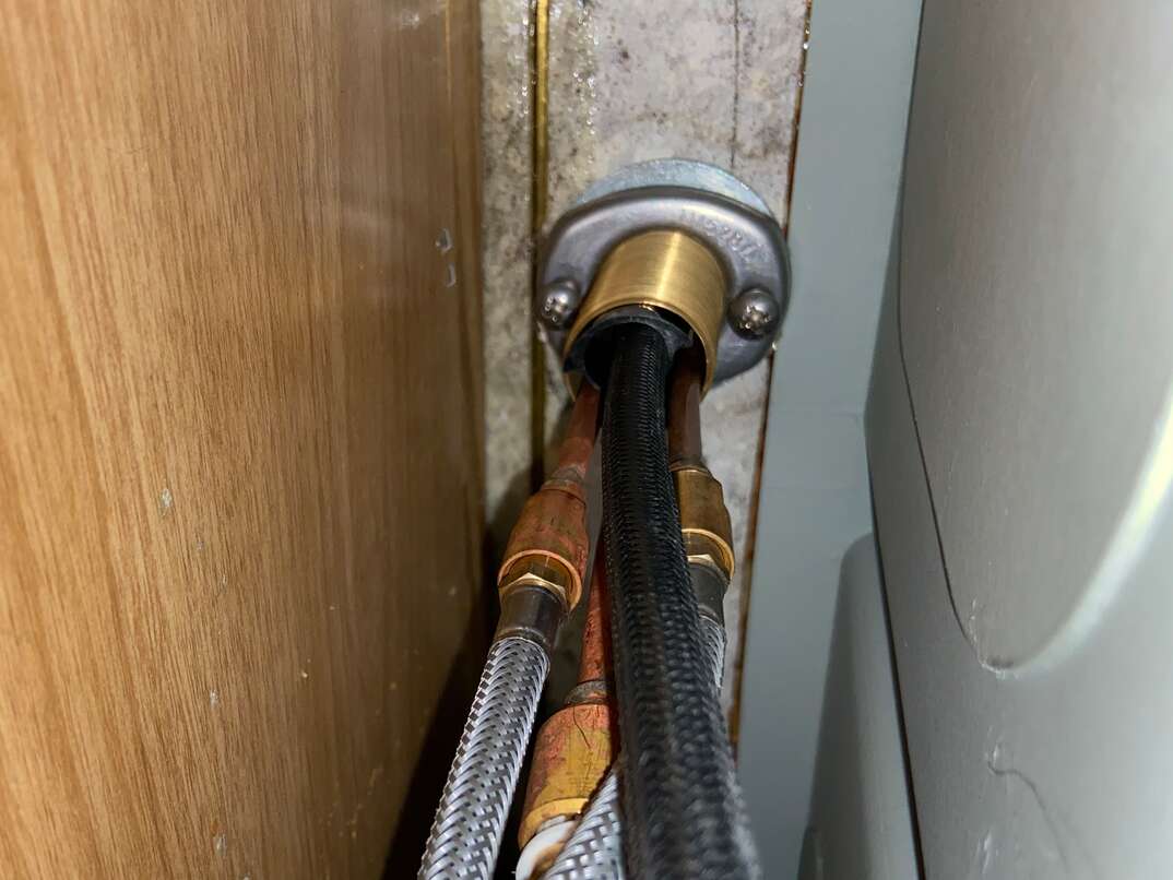 A real photo taken from inside a kitchen sink cabinet showing the underside of a single-handle faucet with two braided supply lines and a mounting nut