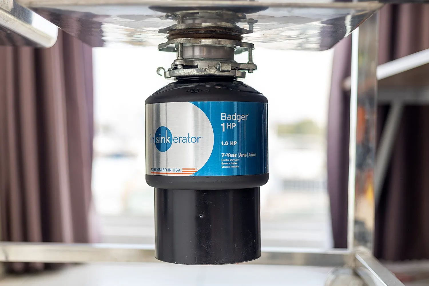 A stainless steel garbage disposal installed under a kitchen sink with the mounting ring and drain connections visible