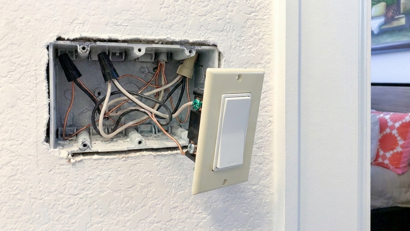 A standard bathroom fan switch pulled out from the wall box, showing black and white insulated conductors and a bare copper ground wire