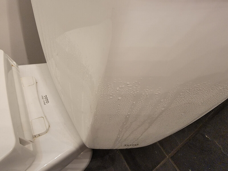 A standard white toilet tank with visible condensation droplets beaded on the outside porcelain surface in a humid bathroom