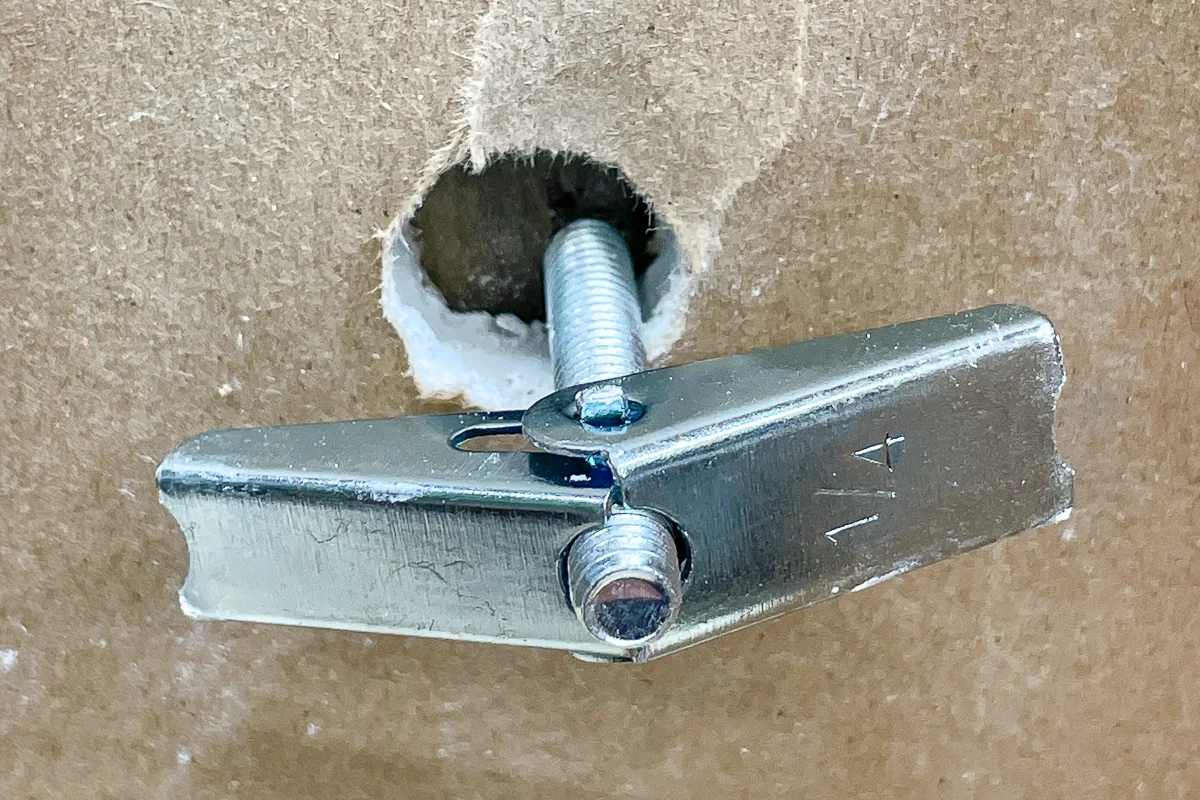 A toggle bolt being inserted through a drilled hole in drywall with the toggle opened behind the wall and the screw being tightened
