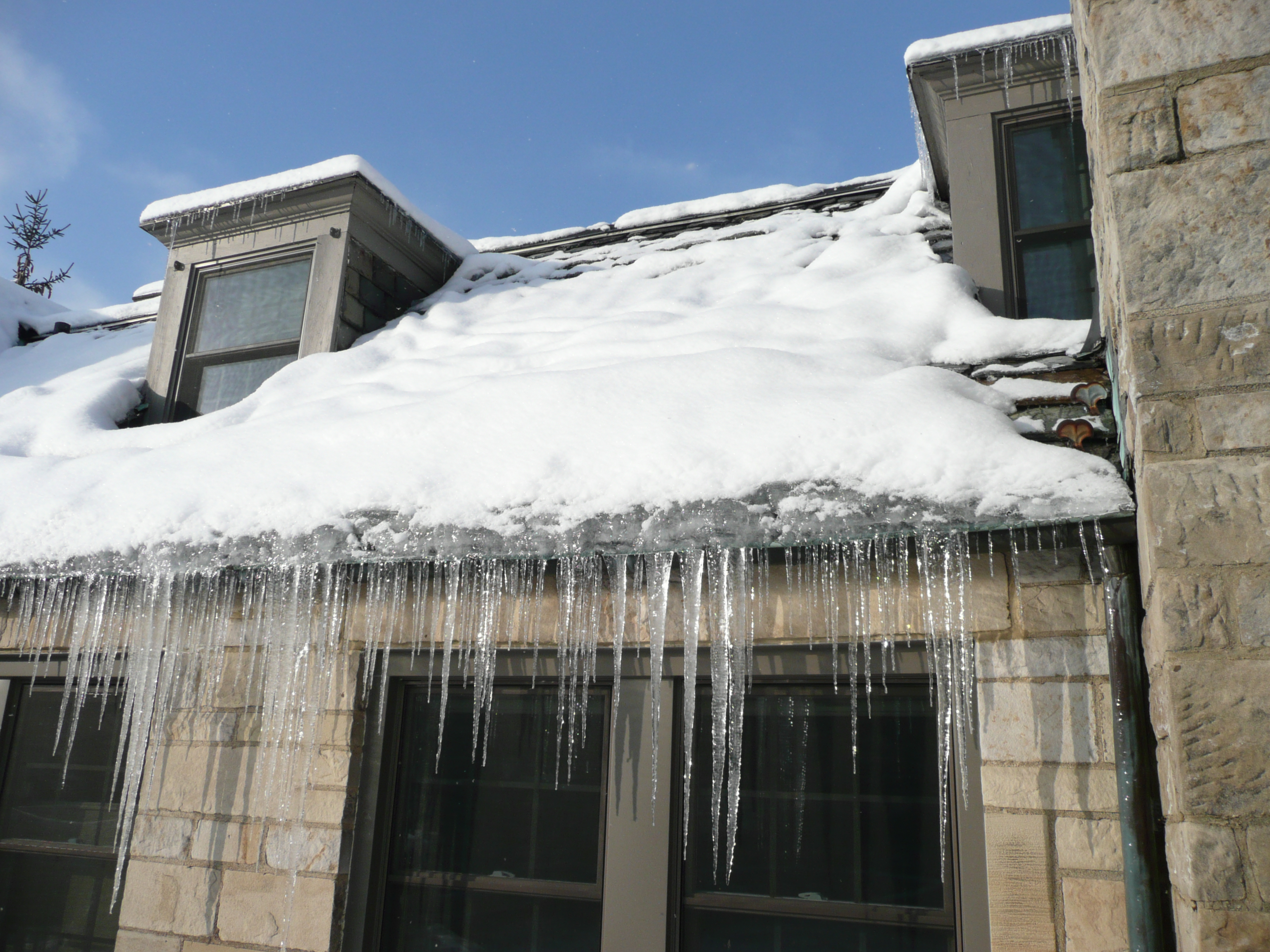 A winter roof edge with a thick ice dam and long icicles hanging from the eaves above a snowy yard