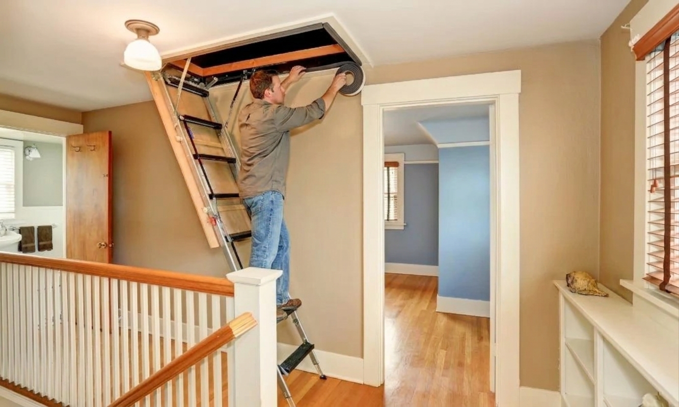 Adhesive foam weatherstripping being applied along the top edge of an attic stair frame on the ceiling side