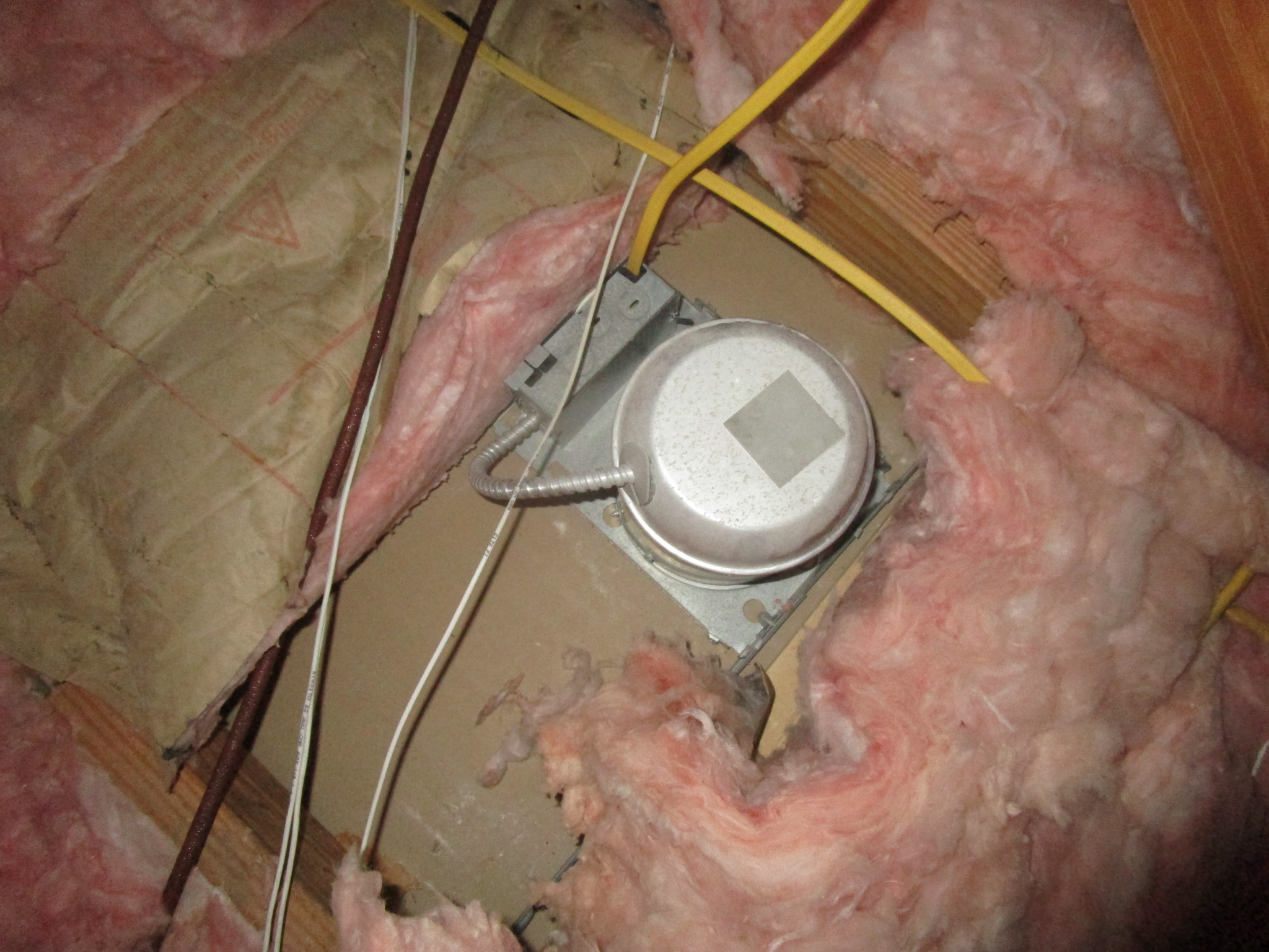 An open attic showing a recessed can light housing protruding above the ceiling drywall with loose-fill insulation nearby in a real home attic