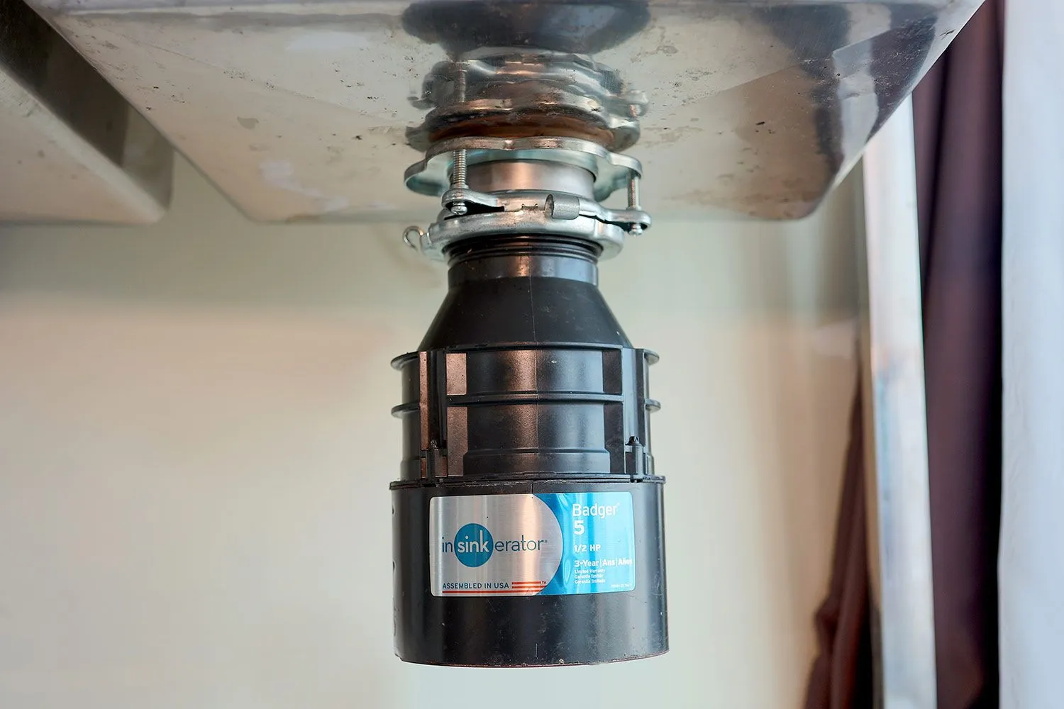 An under-sink garbage disposal mounted to a kitchen sink drain with the electrical cord unplugged and the reset button visible on the bottom