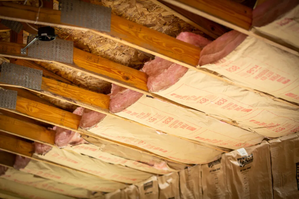 An unfinished attic floor with existing fiberglass batt insulation between joists, a few areas of loose-fill insulation, and exposed framing, photographed from the attic access opening