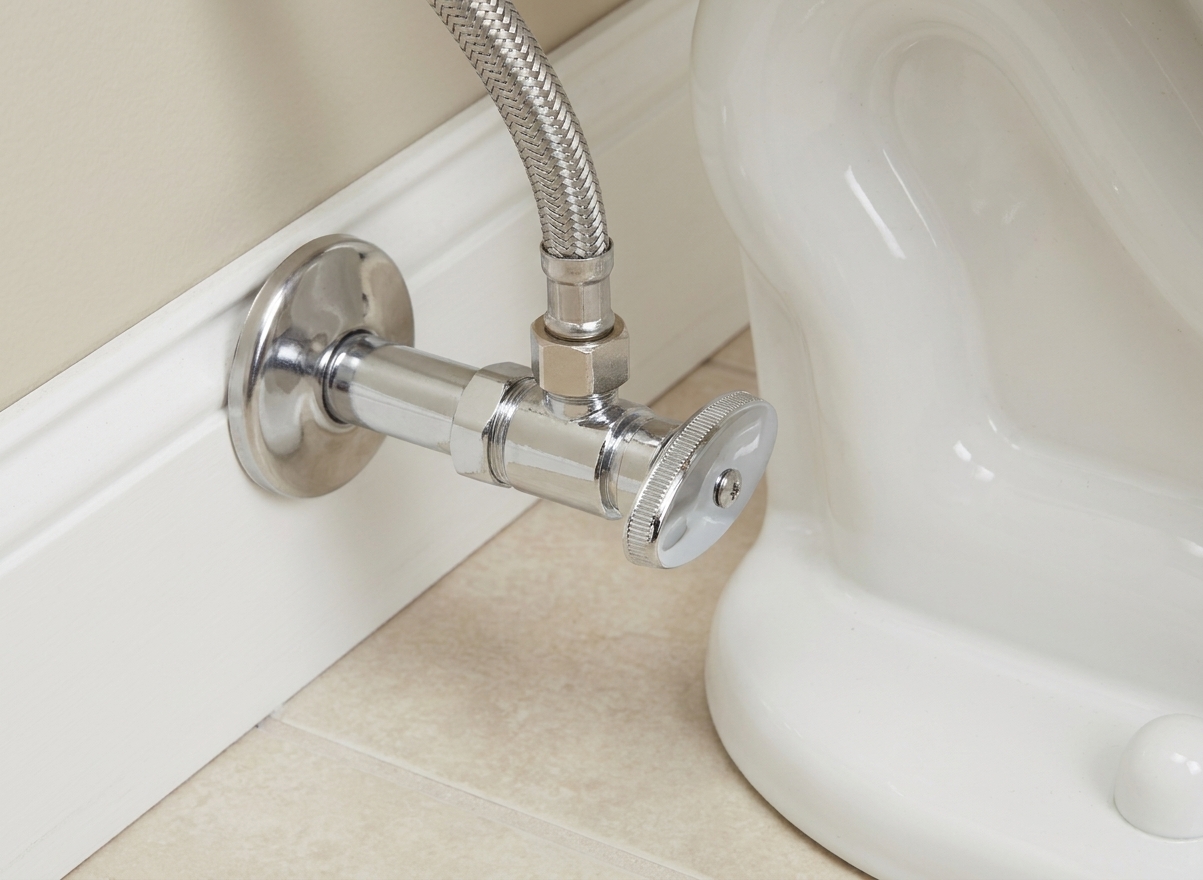 Close-up photo of a chrome angle stop shutoff valve behind a toilet with a braided supply line nut attached