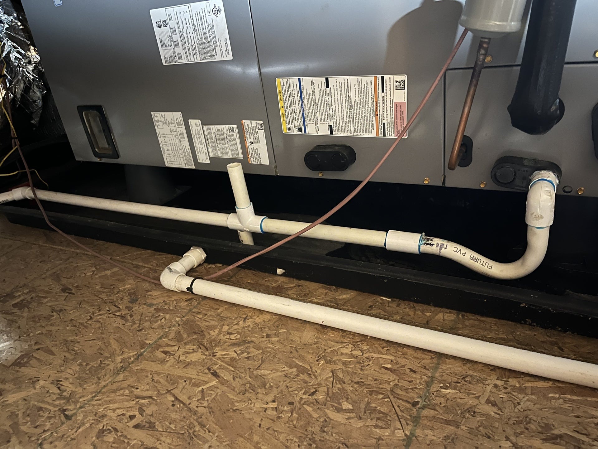 Closeup photo of a white PVC condensate drain line coming off an indoor air handler in a utility closet, with fittings and a visible cleanout tee