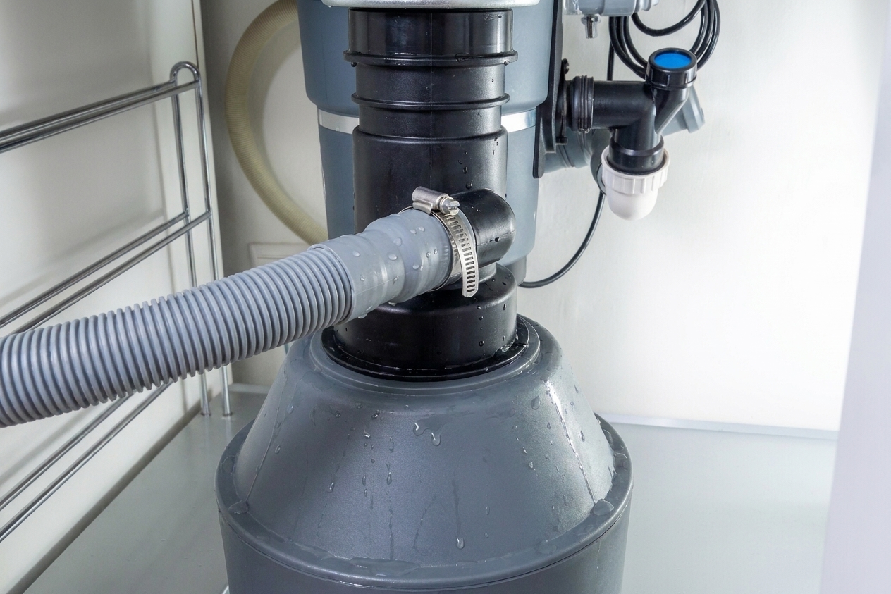 Closeup under-sink photo of a dishwasher drain hose attached to a garbage disposal inlet with a metal hose clamp