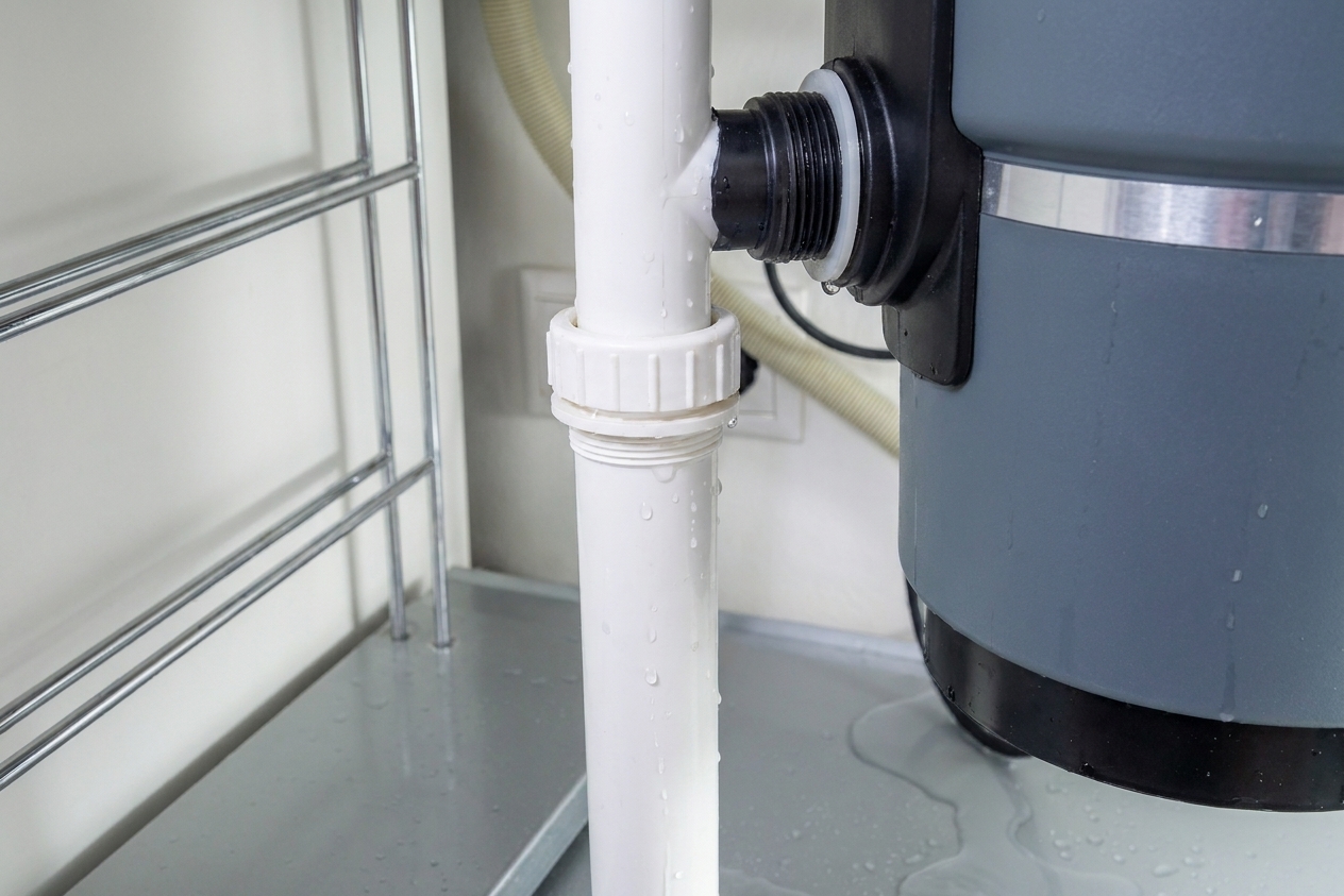 Closeup under-sink photo of the garbage disposal discharge outlet connected to a white drain pipe with a slip nut and washer